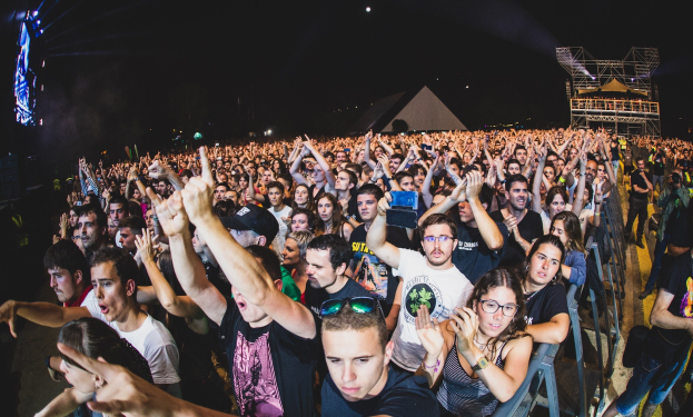 Menschenmenge auf einem Musikfestival mit erhobenen Händen, einige sitzen auf Stühlen und andere stehen, in der Nähe eines Metallzauns mit Gebäuden und Lichtern im Hintergrund unter einem dunklen Himmel.