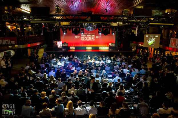 Eine große Menschenmenge sitzt auf Stühlen vor einer Bühne mit Bildschirmen und Bannern, die von Scheinwerfern beleuchtet wird und von Geländern umgeben ist, und blickt erwartungsvoll auf ein Online-Marketing-Rockstars-Event, mit einem Wasserzeichen in der linken unteren Ecke.