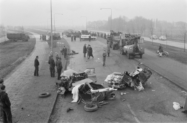 Schwarz-weißes Bild eines Autounfalls am Straßenrand mit mehreren Fahrzeugen, einer Gruppe von Menschen in der Nähe, Laternenpfählen, Bäumen und dem Himmel im Hintergrund.