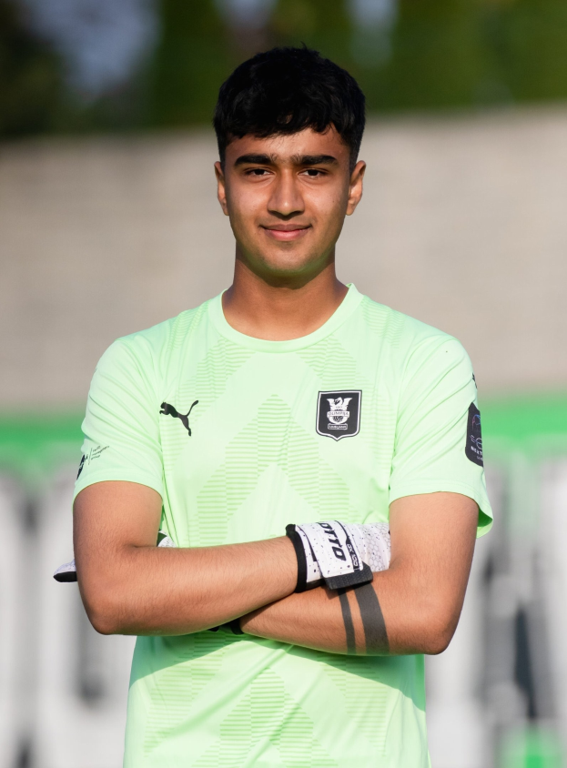 Ein junger Mann in einem grünen Hemd steht auf einem Fußballfeld, die Arme verschränkt und lachend, mit einem unscharfen Hintergrund.