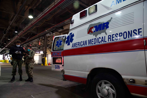 Ein Notarztwagen der American Medical Response (AMR) steht in einem Gebäude geparkt, mit zwei maskierten Personen davor, einem Fahrzeug auf der linken Seite und mehreren Personen, Lichtern und Rohren im Hintergrund.