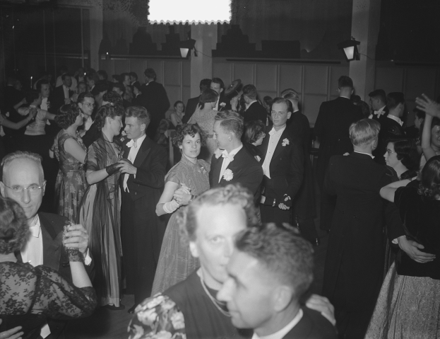 Gruppe von Menschen, die auf einer Party in einem Schwarz-Wei├č-Setting tanzen, mit M├Ąnnern und Frauen verschiedenen Alters, einige halten Gl├Ąser, und eine beleuchtete Wand im Hintergrund.