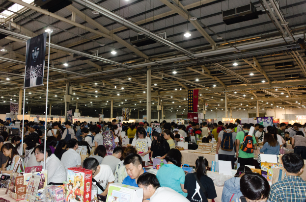 Eine große Gruppe von Menschen umringt Tische voller Bücher, Kartons und anderer Gegenstände auf einer Buchmesse, mit Bannern und Lichtern darüber.