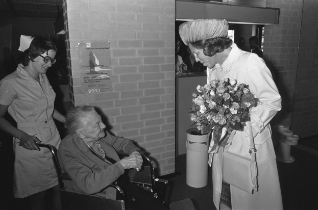 Eine ältere Frau im Rollstuhl, die einen Blumenstrauß hält, wird von einer lächelnden Krankenschwester begrüßt, mit einem Plakat, Mülltonne und anderen Menschen im Hintergrund, alles in Schwarz-Weiß dargestellt.