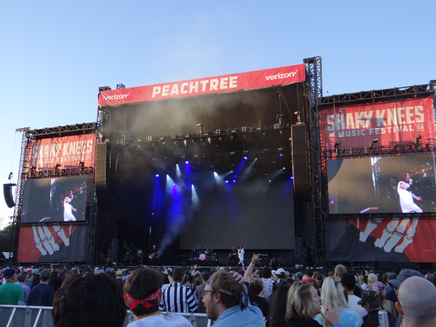 Eine große Menschenmenge steht vor einer hell beleuchteten Bühne auf dem Shaky Knees Music Festival, auf der Musiker mit Instrumenten und Bannern hinter ihnen spielen, unter einem klaren blauen Himmel.