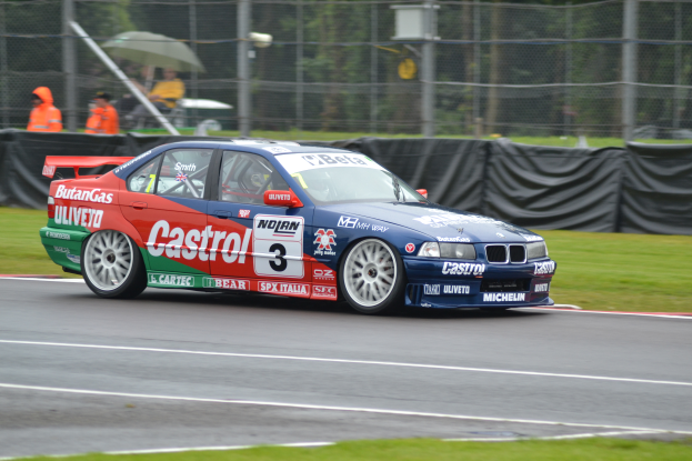 Ein Auto fährt auf einer Rennstrecke mit Gras auf beiden Seiten, mit einem Maschendrahtzaun, Menschen in orangefarbenen Jacken mit Schirmen und Bäumen im Hintergrund.