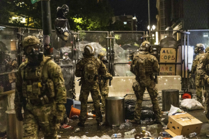 Eine Gruppe von Polizisten in Uniformen und Helmen steht in einer Reihe bei Nacht, hält Gewehre und hat Müll und Trümmer auf dem Boden sowie Gebäude, Bäume und ein Schild im Hintergrund.