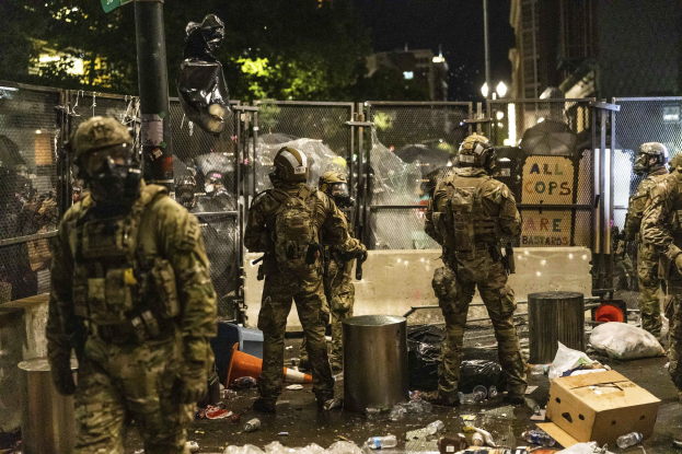 Eine Gruppe von Polizisten in Uniformen und Helmen steht in einer Reihe bei Nacht, hält Gewehre und hat Müll und Trümmer auf dem Boden sowie Gebäude, Bäume und ein Schild im Hintergrund.