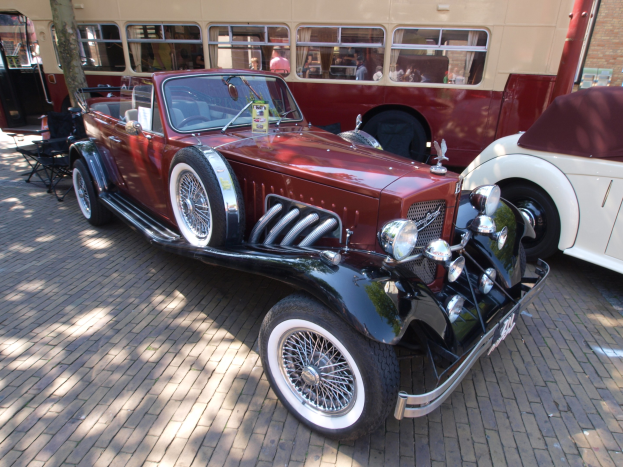 Vintage-Auto neben einem Doppeldeckerbus auf einem Kopfsteinpflasterweg geparkt, mit Stühlen und einem Baum im Vordergrund, einem Gebäude mit Fenstern im Hintergrund und Menschen im Bus zu sehen.