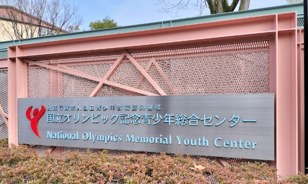 Außenansicht des Nationalen Olympischen Memorial Youth Center mit einem Namensschild, Pflanzen, Bäumen, Gebäuden und einem klaren blauen Himmel.