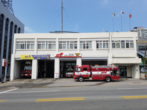 Feuerwache mit mehreren Feuerwehrautos vor dem Gebäude geparkt, Gebäude mit Fenstern, Bannern und Flaggen, bewölkter Himmel und ein Turm auf der linken Seite.