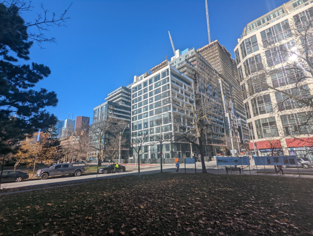 Eine Straße in Toronto mit hohen Gebäuden, fahrenden Fahrzeugen, Passanten auf dem Gehweg, Bäumen und verstreuten trockenen Blättern auf dem Boden unter einem sichtbaren Himmel.