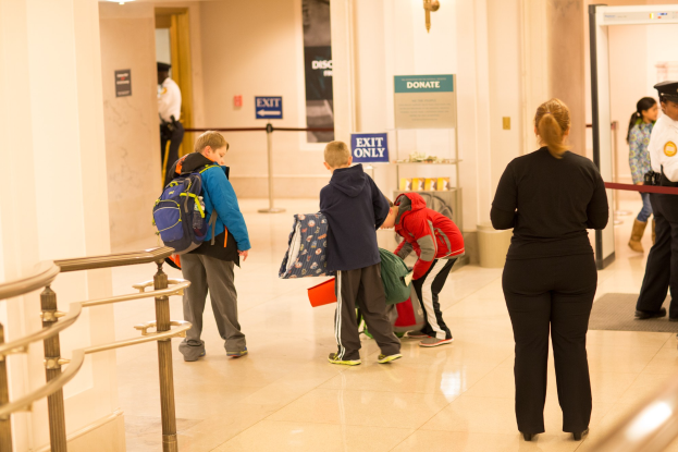 Eine Gruppe von Menschen steht in einem Flur mit einem Geländer auf der linken Seite und Deckenleuchten, mit Tafeln im Hintergrund, die den ersten Schultag im U.S. Capitol anzeigen.