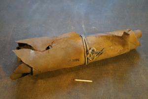 Eine braune Papiertüte, die wie eine Granate aussieht, mit einem Griff, zugebunden mit einem Seil und eine Zigarette daneben auf einem Tisch.