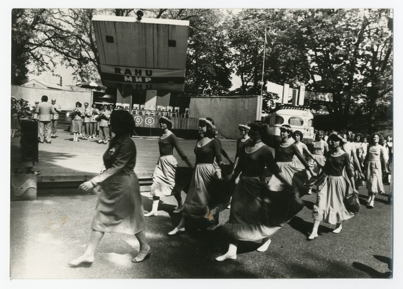 Eine Gruppe von Frauen, die auf der Straße tanzen, umgeben von einer Menge, einem Lautsprecher, einem Fahrzeug, einer Tafel mit Text und einem Fahnenmast.