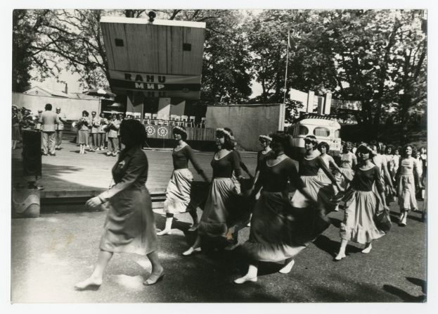Eine Gruppe von Frauen, die auf der Straße tanzen, umgeben von einer Menge, einem Lautsprecher, einem Fahrzeug, einer Tafel mit Text und einem Fahnenmast.