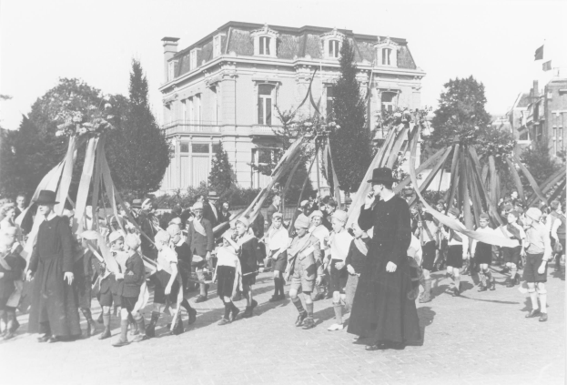 Schwarz-weißes Bild einer Gruppe von Menschen, die eine Straße entlanggehen und Fahnen schwenken, mit Hüten, vor Gebäuden und Bäumen, mit einem Maibaum im Hintergrund.