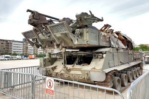 Ein gepanzerter Transporter vor einem Metallzaun mit einem daran befestigten Schild, mit Fahrzeugen, Bäumen, Gebäuden und einem klaren blauen Himmel im Hintergrund.