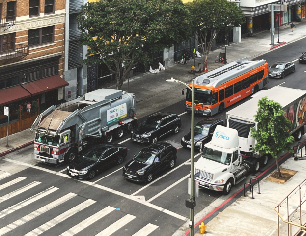 Stadtstraße mit starkem Verkehr von Autos, Bussen und Lastwagen, gesäumt von Bäumen, Laternenmasten und Gebäuden mit Fenstern, mit Geländern, Treppen, Fußgängern auf dem Gehweg und Texttafeln.