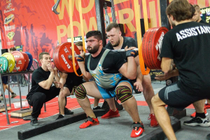 Eine Gruppe von Männern in einem Fitnessstudio, die Kniebeugen mit Hanteln machen, umgeben von Fitnessgeräten und Bannern mit der Aufschrift "Powerlifting-Weltmeisterschaften 2019".