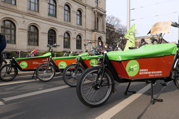 Gruppe von Fahrrädern, die an der Seite einer Straße geparkt sind, mit einer Person in der Nähe, Gebäuden und Bäumen im Hintergrund bei klarem blauem Himmel und einer Tafel, die eine Fahrrad-Sharing-Kampagne anzeigt.