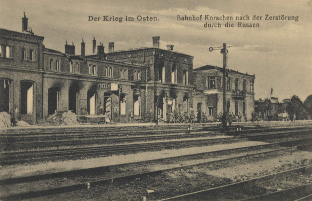 Schwarzes und weißes Foto des Bahnhofs Korschen in Ostern, Deutschland, mit Schienensträngen, Gebäuden, Strommasten, Menschen, Bäumen und einem Himmel-Hintergrund mit Text oben und unten.
