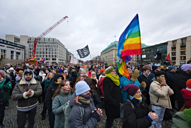 Eine große Gruppe von Menschen mit Fahnen und Schildern vor einem Gebäude während einer LGBTQ+ Demonstrationen in Berlin, mit Gebäuden, einem Kran und Wolken im Hintergrund.