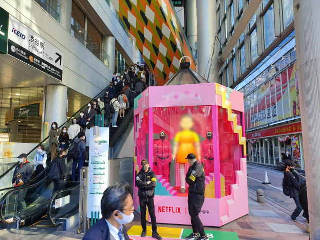 Eine Gruppe von Menschen steht vor einem gläsernen Gebäude mit Säulen und Texttafeln, einige tragen Masken, während hinter ihnen eine Rolltreppe mit Menschen fährt; ein pinker Kasten in der Mitte zeigt Schaufensterpuppen und trägt die Aufschrift "Netflix's neuer Netflix-Stand in Tokyo."