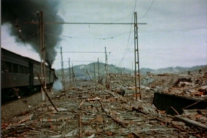 Ein Zug fährt durch ein zerstörtes Gebiet mit Rauch, der aus ihm aufsteigt, umgeben von Strommasten und -leitungen, mit Hügeln und einem klaren Himmel im Hintergrund und verstreuten Trümmern am Boden.