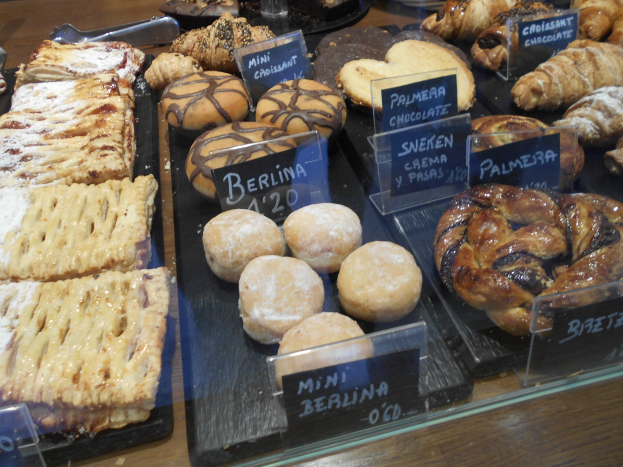 Ein Holztisch mit einer Vitrine voller verschiedener Gebäcke, darunter Brötchen und Kuchen, sowie Namensschilder, die auf eine Bäckerei hindeuten.