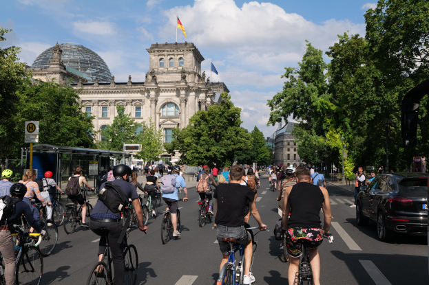 Eine Gruppe von Menschen, die auf Fahrrädern eine von Bäumen gesäumte Straße in Berlin, Deutschland, entlangfährt, mit einer Bushaltestelle auf der rechten Seite und einer Flagge, die auf einem Gebäude weht, unter einem bewölkten Himmel.