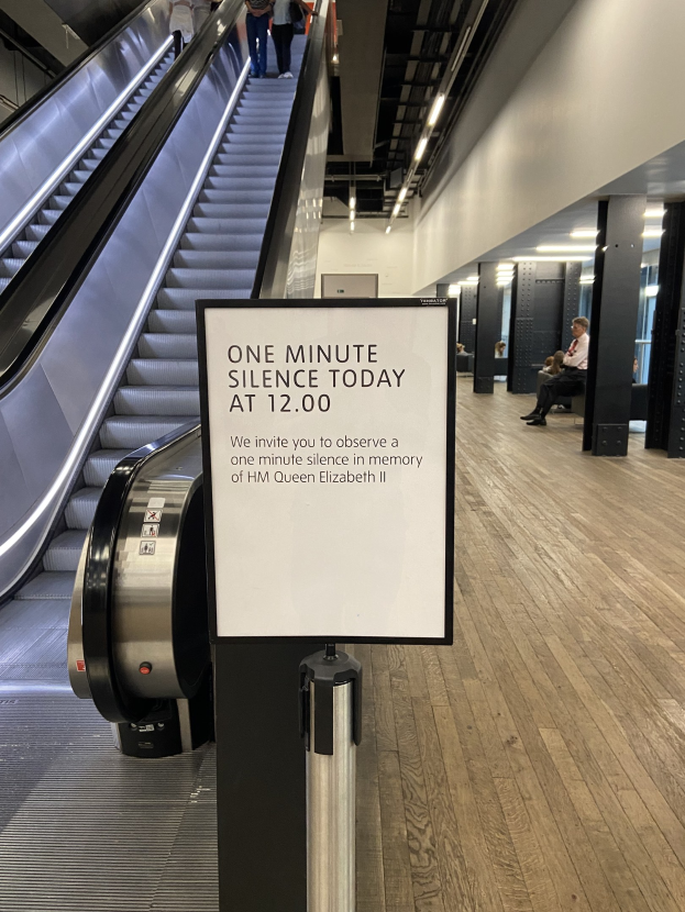 Eine Rolltreppe in einem Flughafen mit einer Tafel, auf der "Eine Minute Stille heute" steht, einige Menschen darauf und an der Decke angebrachte Lampen im Hintergrund.