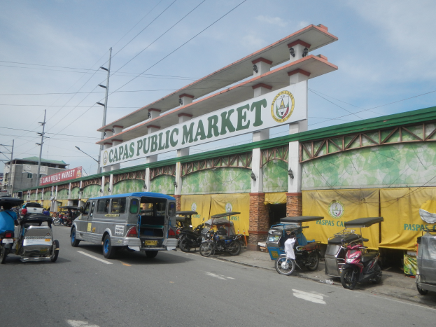 Eine belebte Stadtstraße mit Autos, Motorrädern und Rikschas vor einem Gebäude mit der Aufschrift 'Capas Public Market', umgeben von Strommasten, Laternenmasten und einem bewölkten Himmel.