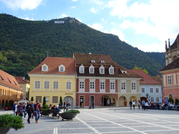 Eine Gruppe von Menschen, die durch einen Stadtplatz mit Gebäuden, Fenstern, Laternenmasten und Topfpflanzen geht, mit einem Hügel voller Bäume und einem bewölkten Himmel im Hintergrund.