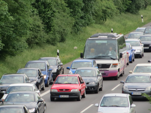 Verkehrsstau auf einer Autobahn mit mehreren Autos und einem Lieferwagen, Menschen in den Fahrzeugen, Büsche und Gras im Hintergrund.