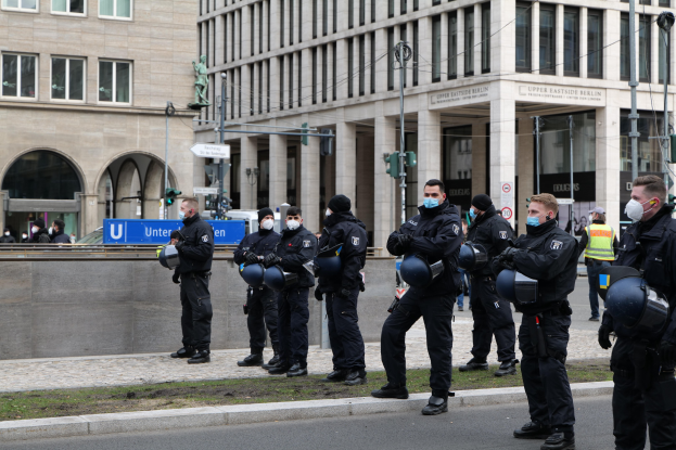 Gruppe von Polizisten in schwarzen Uniformen und Masken vor einem Gebäude in Berlin, Deutschland, mit Glasfenstern und Säulen, Laternen, Verkehrszeichen, Schildern mit Text und anderen Menschen im Hintergrund.