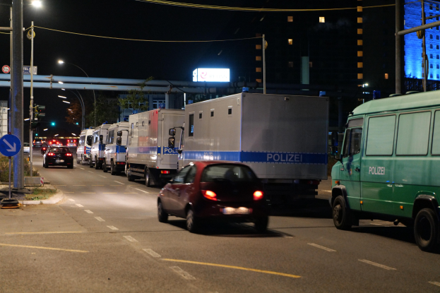 Lange Reihe von Polizeiwagen und -lastwagen, die nachts am Straßenrand geparkt sind, mit Fahrzeugen auf der Straße, Schildern, Pfählen, Laternen, Drähten, Bäumen, Gebäuden und einer Brücke im Hintergrund unter einem dunklen Himmel.