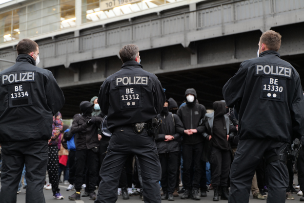 Polizisten in schwarzen Uniformen und Masken vor einer Menge während einer Demonstration, mit einer Brücke und einem Gebäude im Hintergrund.