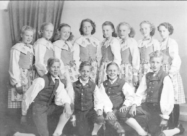 Schwarzes und weißes Foto von Kindern, die vor einer Vorhangwand posieren, mit dem Text "1930er Kinder Schule" unten.