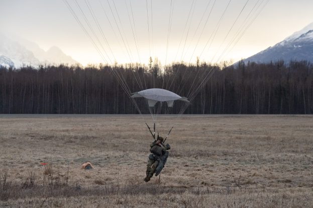 Eine Person beim Paragliding über einer grünen Wiese mit schneebedeckten Bergen im Hintergrund, die einen Helm trägt und gegen einen hellblauen Himmel fliegt.