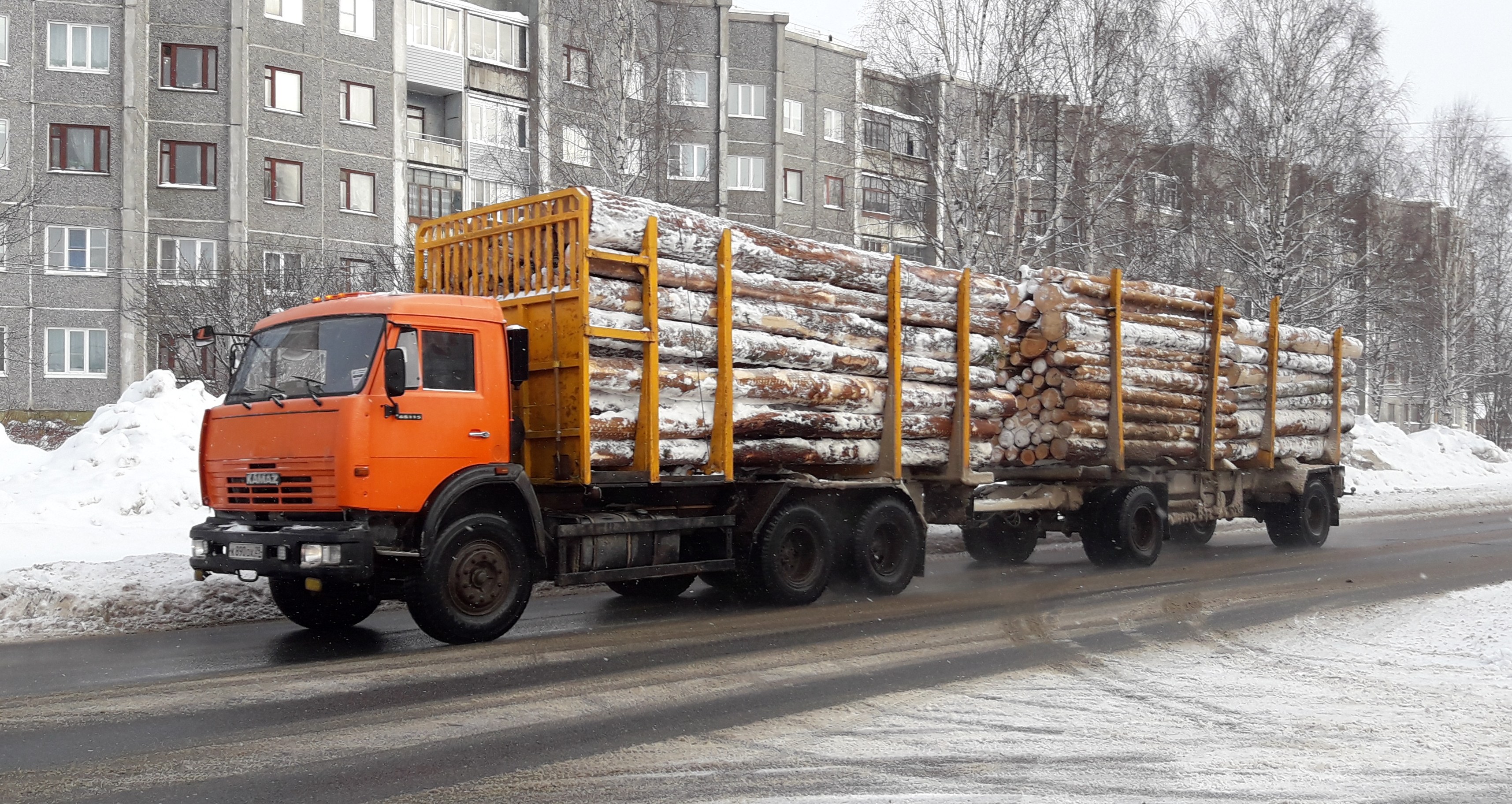 Lastwagen mit Holztransport auf verschneiter Straße mit Bäumen, Fenstergebäuden und einem klaren Himmel im Hintergrund.