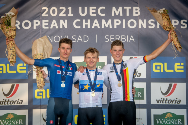 Drei Männer mit Medaillen und Blumensträußen stehen auf einem Podium mit einem Banner "2021 UEC MTB Europameisterschaften" im Hintergrund.