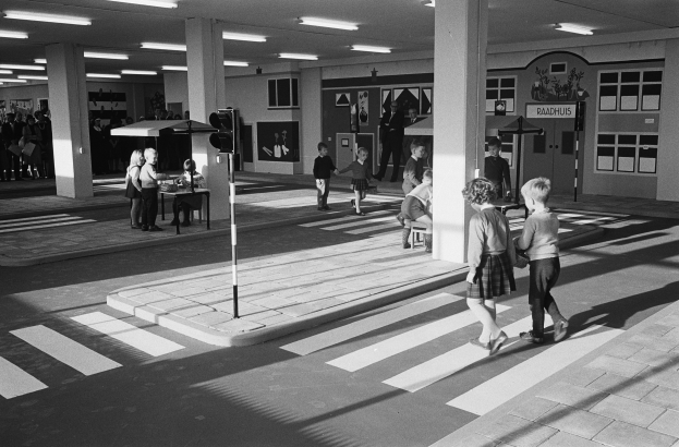 Schwarzes und weißes Foto von Kindern, die eine Straße an einer Ampel überqueren, mit Fußgängern in der Nähe, einer Verkehrsampel im Vordergrund und strukturellen Pfeilern und Deckenleuchten im Hintergrund.