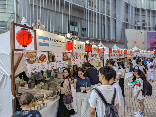 Menschen um einen Tisch mit Essen bei einem Outdoor-Event in Japan, mit hängenden Lichtern und Laternen sowie Informationsschildern in der Nähe.