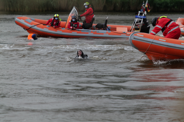 Personen in einem aufblasbaren Boot auf einem Fluss helfen zwei Personen im Wasser, mit Vegetation im Hintergrund; alle tragen Schwimmwesten und Helme.