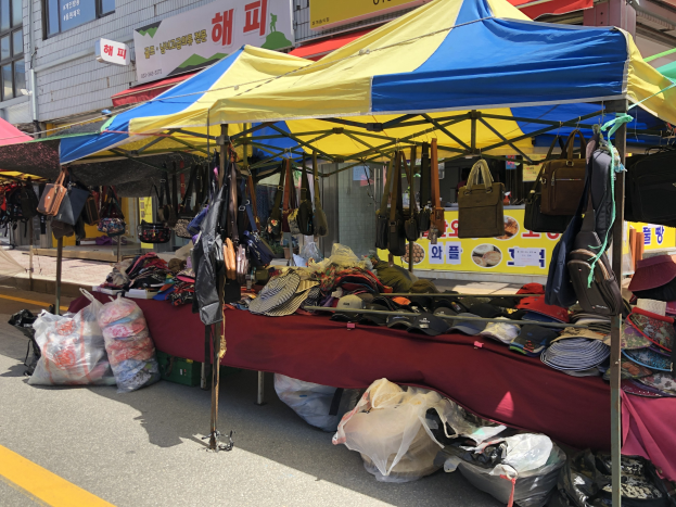 Ein Straßenmarkt mit Zelten, die Taschen, Hüte und andere Gegenstände präsentieren, vor Häusern mit Fenstern und Texttafeln im Hintergrund.
