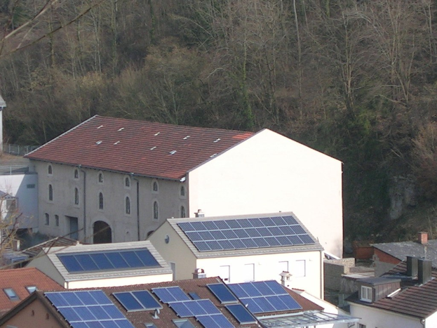 Haus mit Solarpanelen auf dem Dach, umgeben von anderen Gebäuden und Bäumen im Hintergrund.