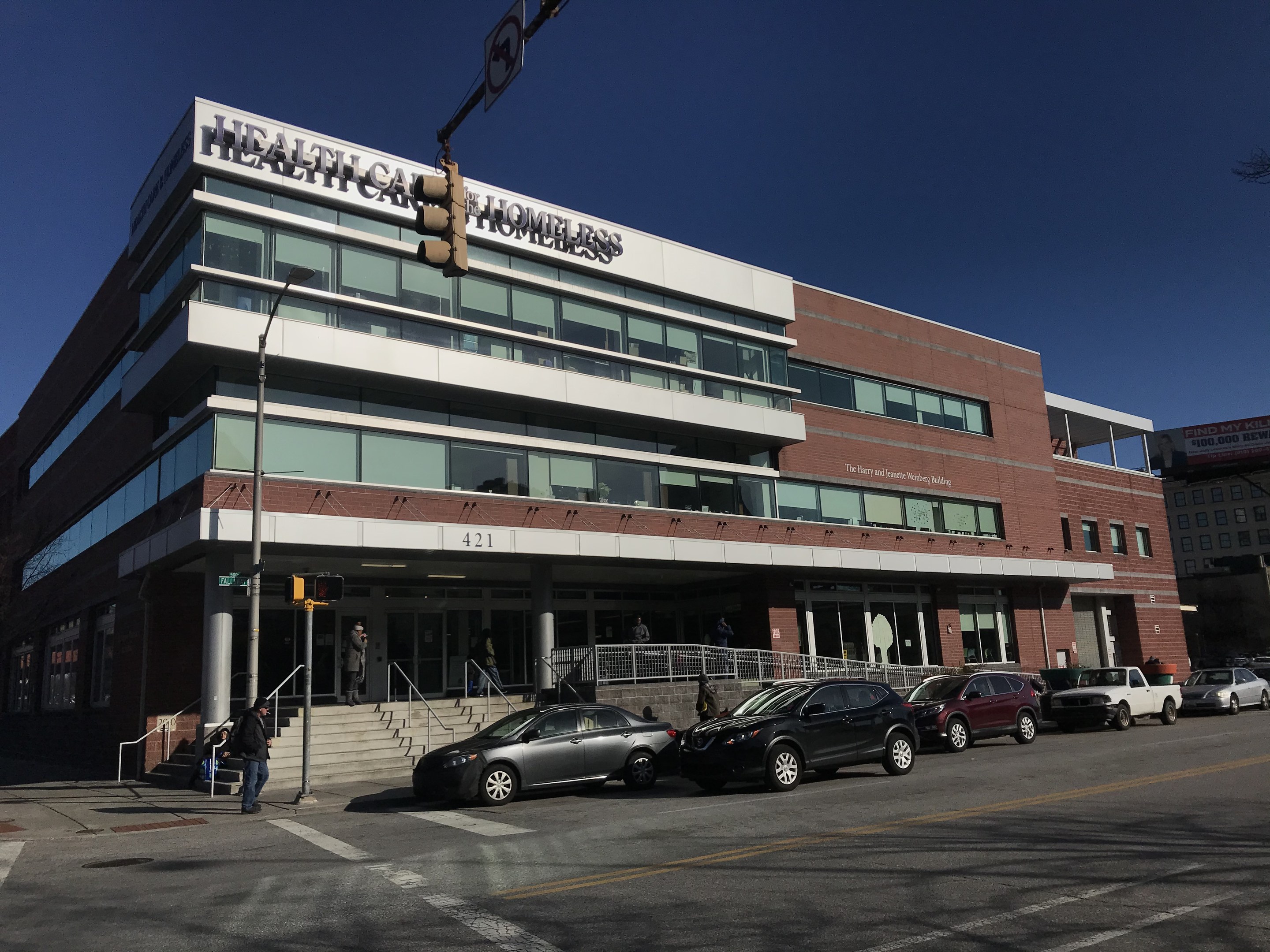 Großes Gebäude mit der Bezeichnung New York City Health Care Homeless Center mit parkenden Autos, Fußgängern, Straßenmöblierung und Bäumen unter einem klaren Himmel.