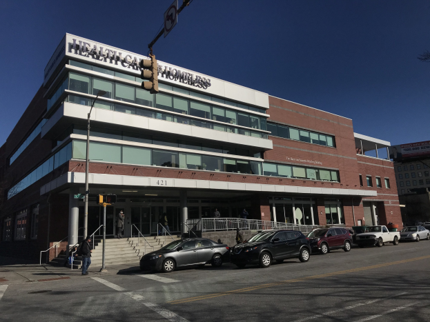 Großes Gebäude mit der Bezeichnung New York City Health Care Homeless Center mit parkenden Autos, Fußgängern, Straßenmöblierung und Bäumen unter einem klaren Himmel.