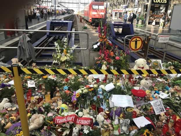 Ein Bahnsteig mit einem Zug im Hintergrund, umgeben von Menschen und verschiedenen Gegenständen wie Teddybären, Blumensträußen und Kerzen, die an einen tragischen Zugunglück in Berlin, Deutschland, erinnern.